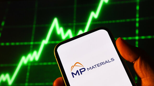 A close up photo of a smartphone screen showing the MP Materials logo with a green financial graph in background.