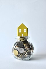 A small yellow wooden house icon on top of a round glass jar filled to the brim with coins, illustrating housing finance, real estate assets, and the accumulation of wealth for property purchases. © Akram