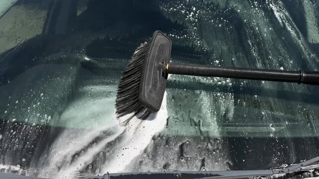 Manual scrubbing of a soapy car windshield using a wash brush with foam.