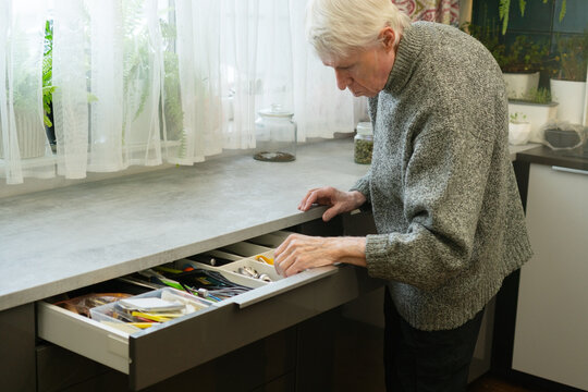 A senior man with white hair opens a drawer filled with cutlery. The image depicts a daily kitchen task, highlighting independence and routine. Elderly person managing household items.
