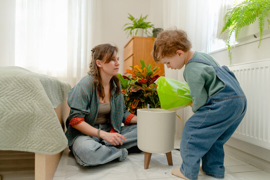A playful mother and son repot a potted plant with colorful foliage on a bright indoor floor. The happy boy holds leaves and helps mom add soil while learning gardening in a cozy living room.
