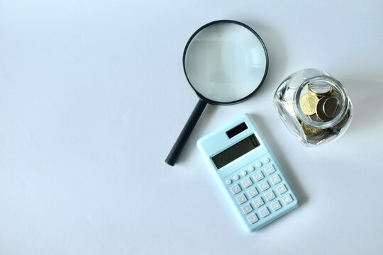 A high-angle studio shot of a black magnifying glass, a light blue digital calculator, and a transparent glass jar filled with coins on a white surface.