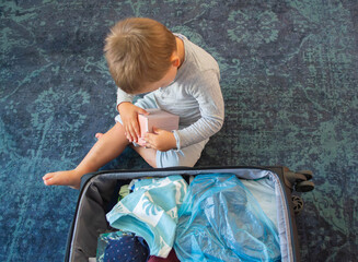 Naklejka premium Cute little boy helping to pack a large black suitcase indoors before a family trip. Toddler playing near open luggage filled with clothes and travel essentials. Highlighting the concept of family .
