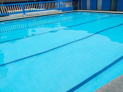 Clean outdoor lap swimming pool with bright blue water and lane lines. Stainless steel pool ladder in foreground, tiled deck and blue walls in background