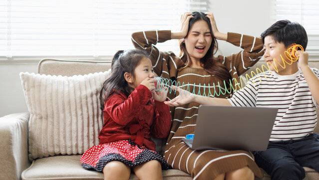 Stressed young mother overwhelmed by noisy playful children using spring toys while working on laptop, experiencing frustration and burnout in living room at home