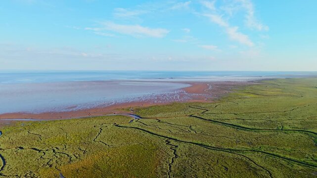 Three keyframes of an aerial coastal landscape shot. Keyframe 1: Aerial view reveals expansive green grassy dunes leading to sandy shore