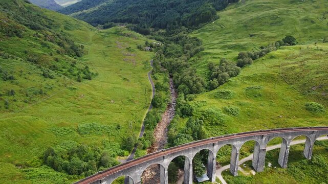 Beautiful aerial of a dry river running under the famous Glenfinnan Viaduct in the highlands of Scotland. The drone flies backwards and reveals a beautiful green valley on a sunny, summer day.