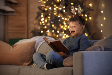 Fototapeta premium Cute little boy reading Christmas story on sofa at home in evening