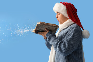 Fototapeta premium Young man in Santa hat and with book blowing snow on blue background