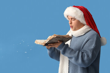 Fototapeta premium Young man in Santa hat and with book blowing snow on blue background