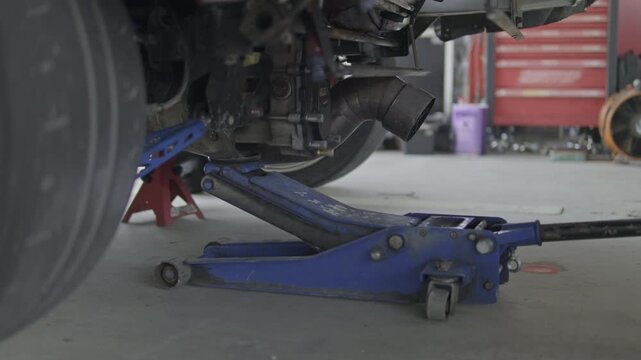 Low-angle close-up shot of a heavy-duty blue hydraulic floor jack lifting the rear subframe and chassis of a track car in a garage.