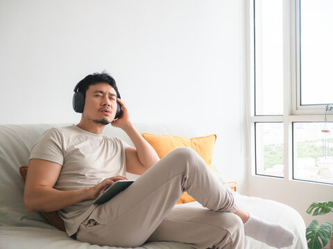 Relaxed Asian man enjoying music with headphones while sitting comfortably on a sofa at home.