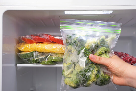 Female hand holds plastic bag with zip fastener with broccoli against the background of an open freezer with other vegetables.
