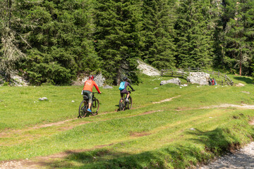 Obraz premium Canazei, Italy. Mountain bikers riding across alpine meadow on forest trail in the Dolomites