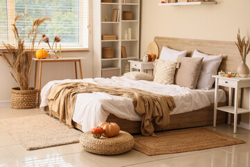 Interior of modern bedroom with comfortable bed, vases of pampas grass, bedside table and pumpkins