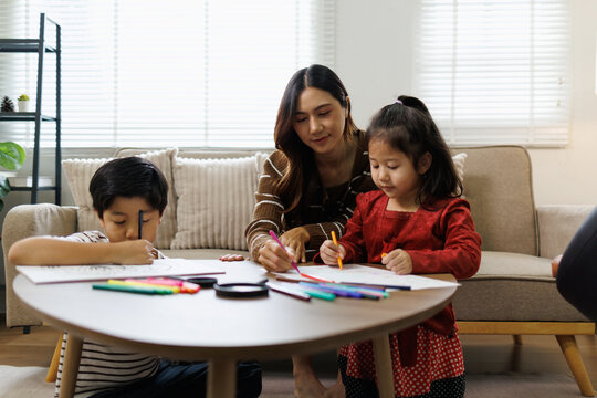 Young asian mother and her two children drawing and coloring together on the living room floor, sharing a creative moment, actively learning and bonding at home