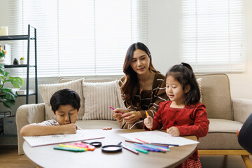 Obraz premium Asian mother and her two young children sitting at a table in the living room, drawing and coloring with markers, engaging in creative playtime and home education