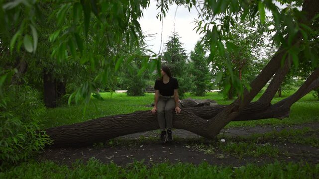 Young woman sitting on fallen tree trunk in shaded park, hands resting on bark, feet dangling, head turned left with thoughtful expression in calm natural setting