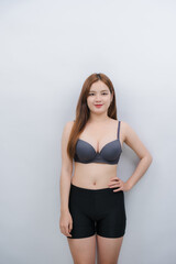 Fototapeta premium Confident Woman Posing in Undergarments: A young woman with long, brown hair stands confidently, wearing a grey bra and black shorts against a light grey background.
