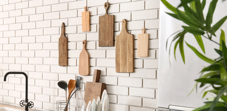 Various wooden cutting boards hanging on a white brick kitchen wall with modern faucet and green plant, a modern black faucet and sink are visible in the foreground