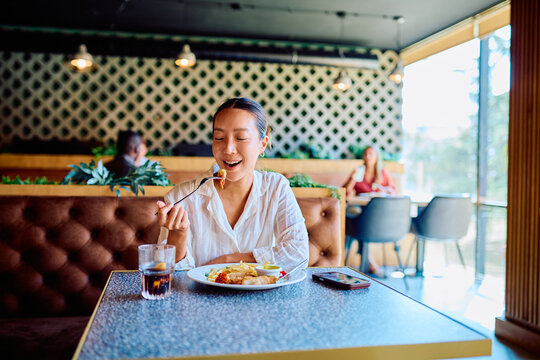 Happy young Asian woman dining alone, savoring a delicious meal at a modern restaurant, eating food with fork