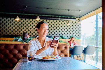Young woman smiling and engaging with her mobile device while enjoying a meal and connectivity in a...