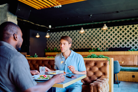 Two multiracial male friends enjoying a casual lunch together at a restaurant, talking and eating at a booth