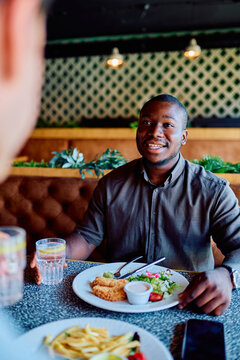 Smiling young African man dining at a restaurant, sharing conversation and eating a healthy meal with a friend