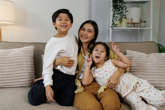 Asian mother embracing son and daughter, family bonding at home, enjoying happy moments on a living room couch, smiling together in a loving and joyful domestic lifestyle scene