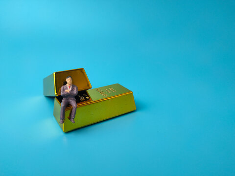 A miniature businessman figure sitting on stacked toy gold bullion bars over a plain blue background