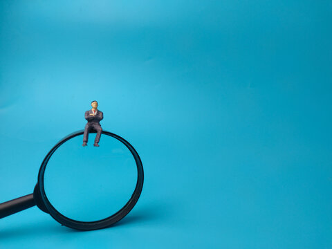 A miniature figurine of a businessman sitting on top of a magnifying glass rim over blue backdrop