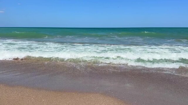Gentle waves roll onto sandy shore under clear blue sky, with white foam and ripples across turquoise water; no text visible