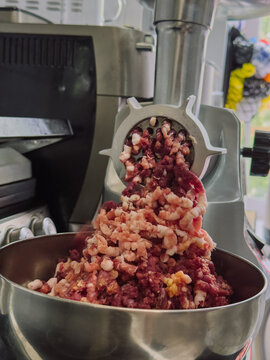 Freshly ground raw meat coming out of electric meat grinder into stainless steel bowl during homemade minced meat preparation.