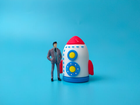 A standing miniature businessman figurine posing next to a colorful toy rocket against a solid blue background