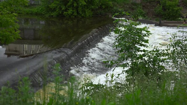 Low concrete weir dividing calm reflective upstream pool from frothy downstream spill, with flowing water, green riverside vegetation, and textured river surface