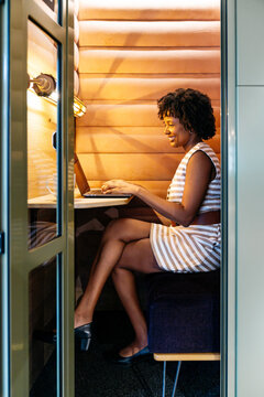 Black woman working on a digital tablet, smiling in a modern office pod, embodying focused work and relaxation