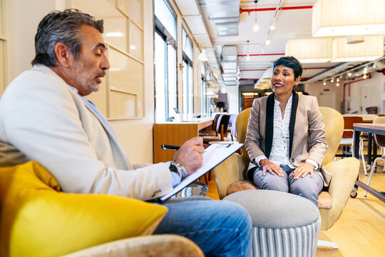 Two business professionals in a relaxed meeting, one taking notes, discussing a project or job interview in a modern office lounge