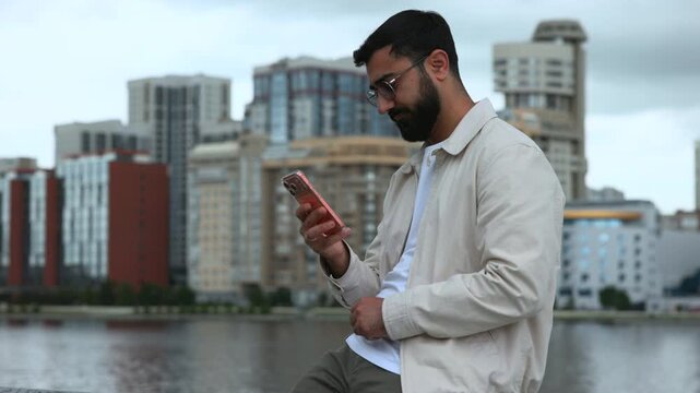 Bearded young man pausing with smartphone near urban waterfront, wearing light beige jacket and looking upward with focused calm under soft overcast city light, with man, bearded, smartphone