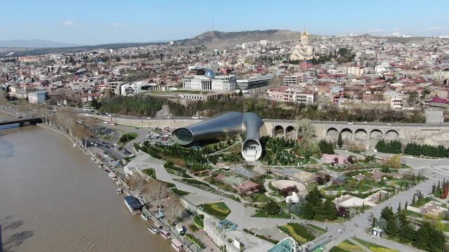 TBILISI, GEORGIA - MAR 10, 2025: Editorial aerial view of Rike Park, the futuristic concert hall, and the Peace Bridge along the river.