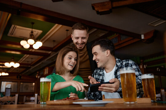 Friends gather around smartphone comparing photos, three adults at wooden table with beers.