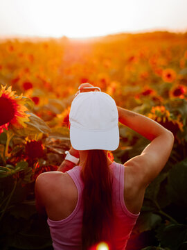 Woman with white baseball cap standing in a vibrant sunflower field during a warm golden sunset