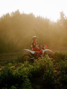 Woman wearing protective gear riding a white dirt bike on a dusty outdoor trail at sunset in summer