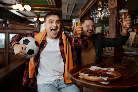 Men cheer with drinks and soccer ball in pub. Joyful men toast during soccer match in cozy bar