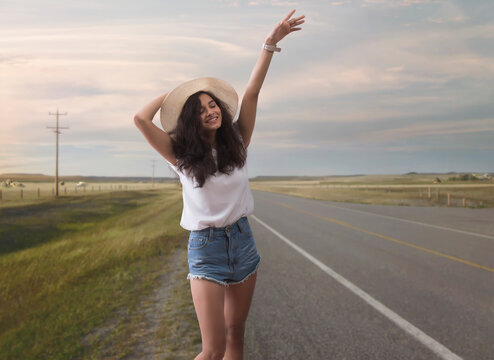 Cheerful woman in hat standing on a deserted highway waving her hand to stop vehicle for a lift