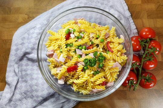 Homemade pasta salad made from fusilli, bell pepper, peas, tomatoes, cooked ham and parsley garnish in a glass bowl on a kitchen towel on a wooden table, top view from above