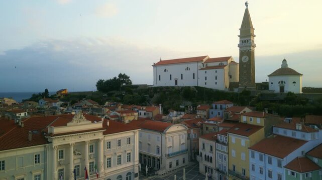 Main square opens in front of church on slope while colorful facades frame plaza at sunset. Clock tower dominates sky near historical buildings in paved center