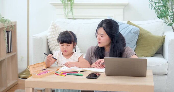 Asian mother watches preschool girl choose color and continue drawing beside laptop on wooden table, supporting early education, problem solving, gentle communication, and close home study routine.