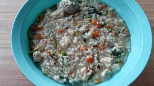 A bowl of warm rice porridge cooked with chopped carrots, green vegetables, tofu, and chicken pieces