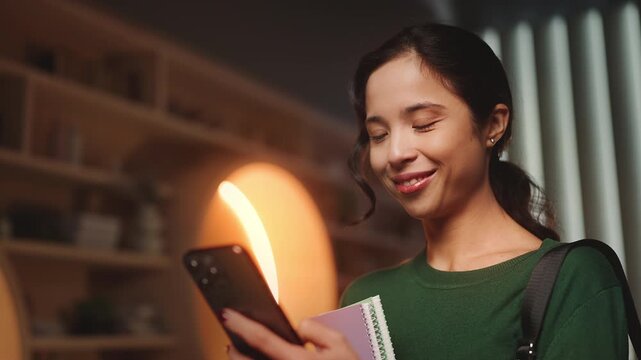 Young Korean woman texting on phone while holding books in library, digital life