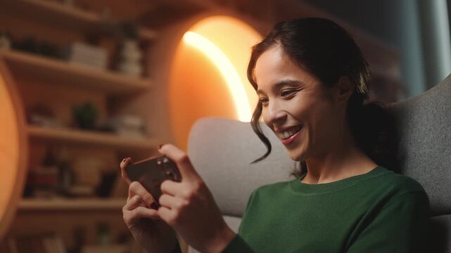 Smiling Asian female playing racing video game on cellphone, home entertainment